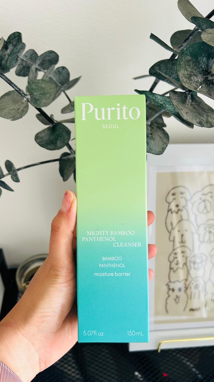 Una mano sostiene una caja de 150 ml de Bemba. PURITO Mighty Bamboo Panthenol Cleanser con extracto de bambú y pantenol, delante de hojas verdes y una ilustración enmarcada de un perro. La caja es verde y azul con texto blanco.
