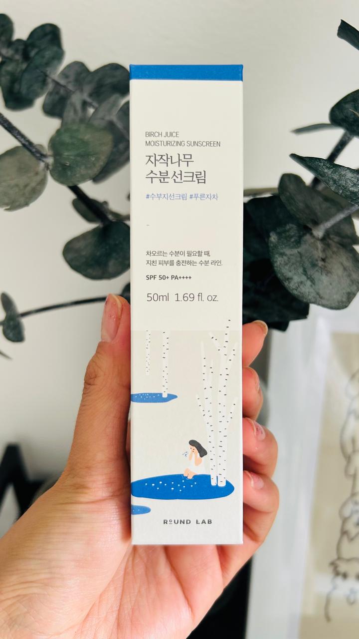 Una mano sostiene una caja de Bemba. Round Lab Birch Juice Moisturizing FPS 50, con texto en coreano y una ilustración de un baño bajo abedules. Este protector solar contiene savia de abedul y niacinamida.