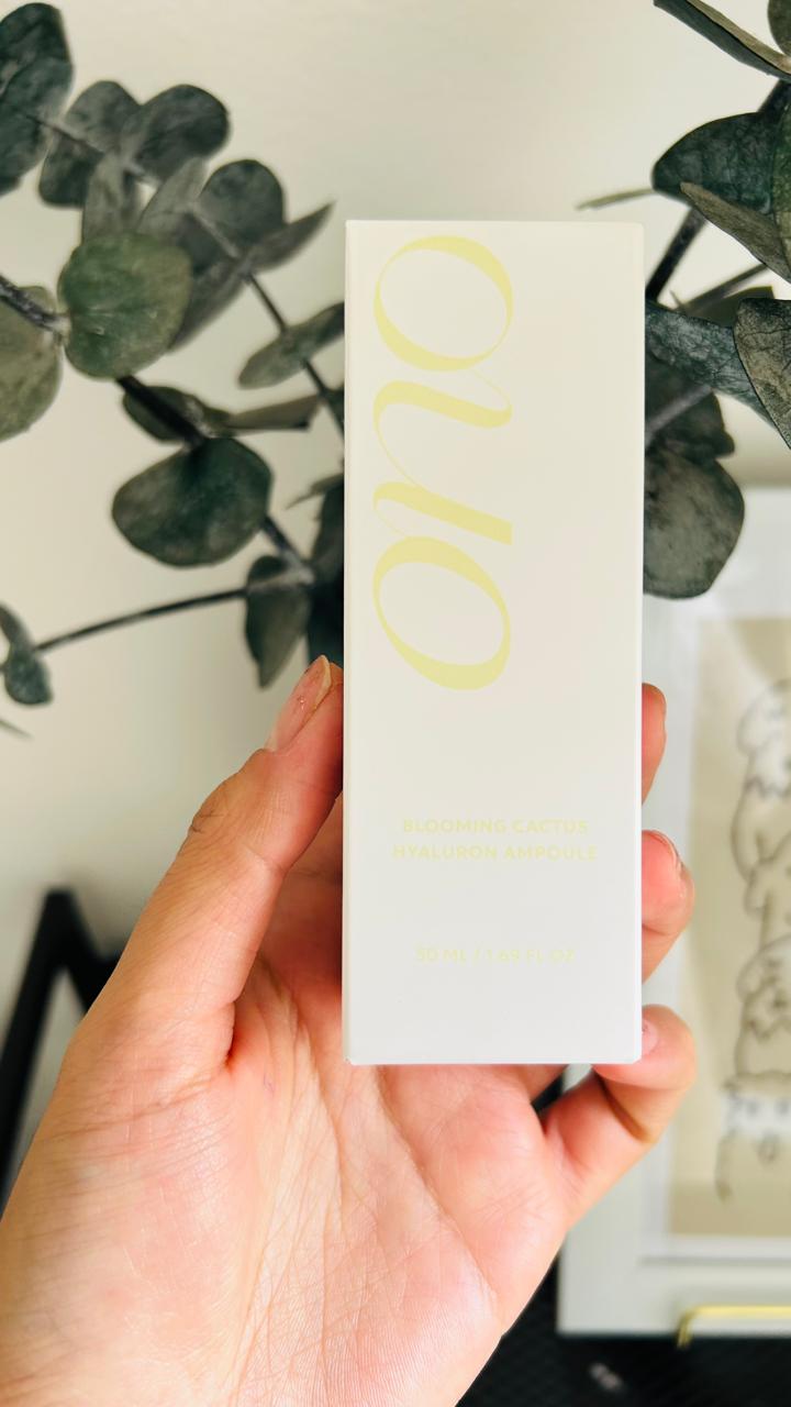 Una mano sostiene una caja rectangular blanca con la etiqueta "Bemba. Onone Blooming Cactus Hyaluronic Ampoule", que ofrece hidratación profunda, con plantas de hojas verdes y obras de arte al fondo.