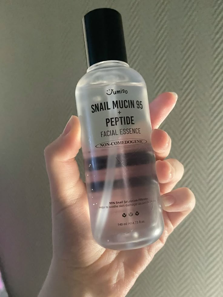 Una mano sostiene un frasco transparente de Bemba. Jumiso Esencia Facial de Mucina de Caracol 95% + Péptidos de 140 ml con tapón negro, que contiene mucina de caracol y péptidos para mejorar la elasticidad de la piel, sobre un fondo texturizado con iluminación tenue.