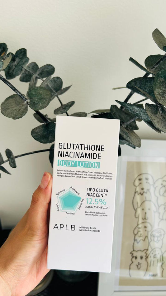 Una mano sostiene una caja blanca de Bemba. APLB Glutathione Loción Corporal, destacando su glutatión y niacinamida para hidratar la piel, frente a hojas verdes y un cuadro con dibujos de gatos al fondo. Los beneficios aparecen en la caja.