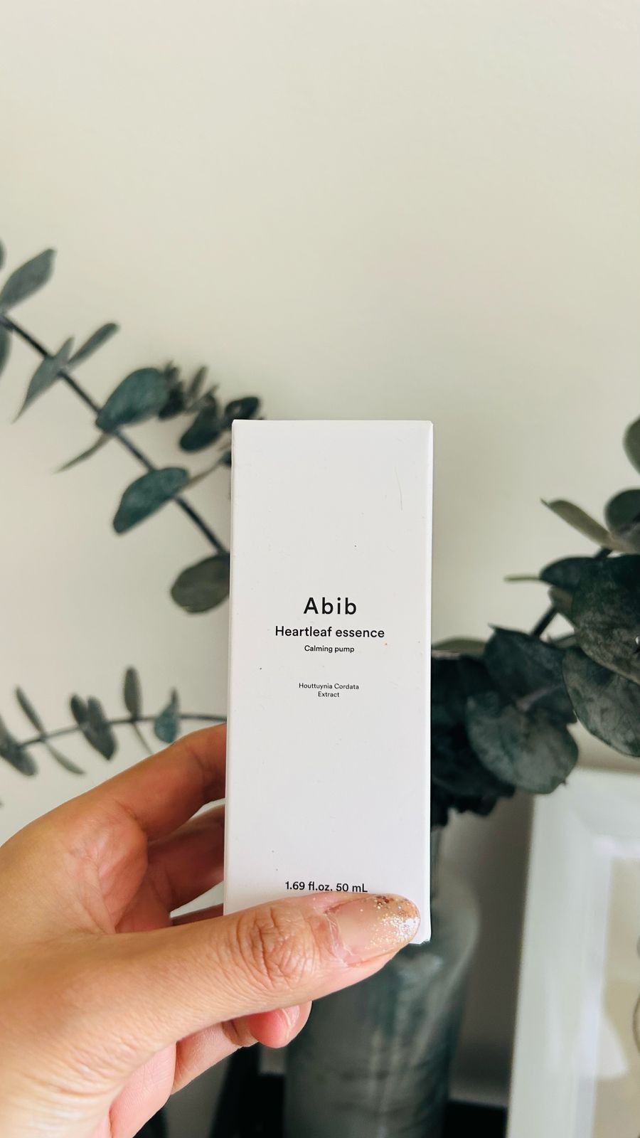 Una mano sostiene una caja blanca de Bemba. ABIB Heartleaf Essence Calming Pump, ideal para pieles sensibles gracias a sus propiedades antiinflamatorias. Al fondo se ve una planta de hojas difusas. La caja contiene 50 ml (1.69 onzas líquidas).