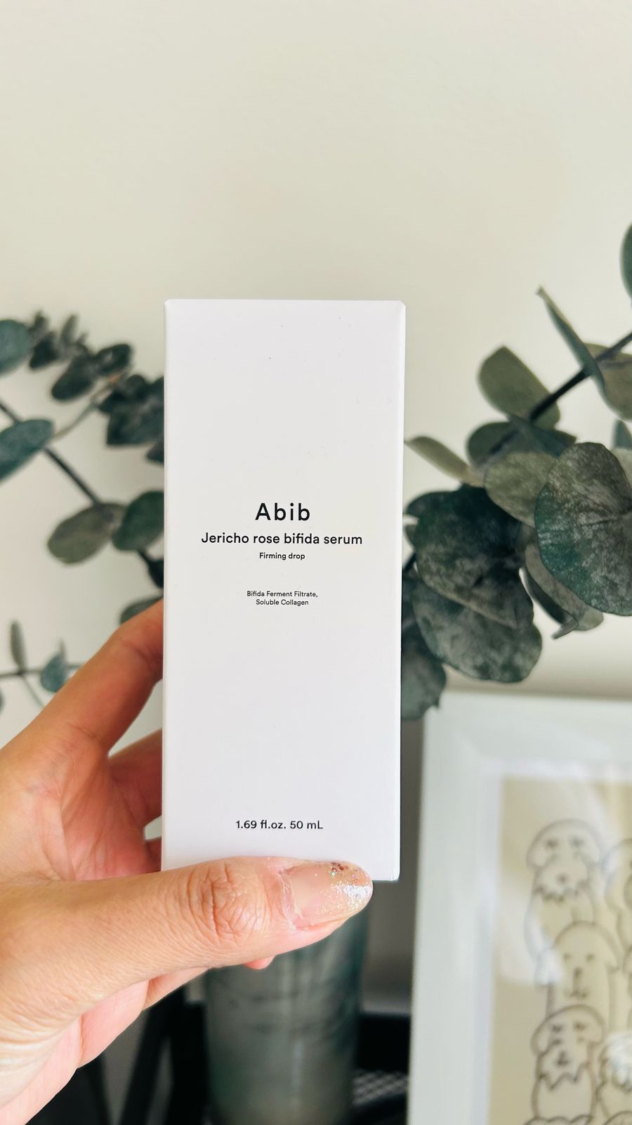 Una mano sostiene una caja blanca de Bemba. ABIB Jericho Rose Essence Bifida, con texto en negro. Hojas de eucalipto y un dibujo enmarcado al fondo. Formulado con Rosa de Jericó para ayudar a fortalecer la barrera cutánea.