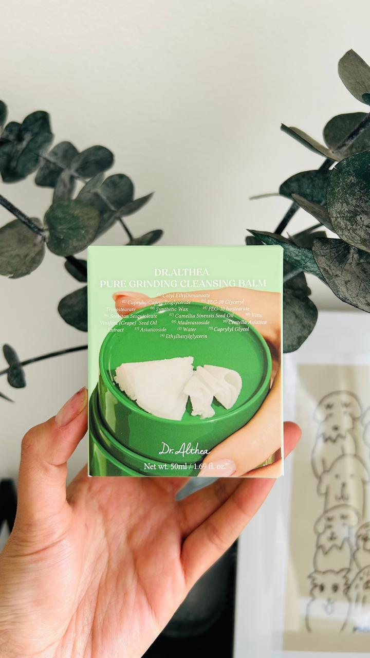 Una mano sostiene una caja verde de Bemba. Bálsamo Limpiador Dr. Althea con centella asiática, con un fondo de ramas frondosas y obras de arte. El envase muestra el bálsamo y la espátula, ideales para eliminar el maquillaje fácilmente.