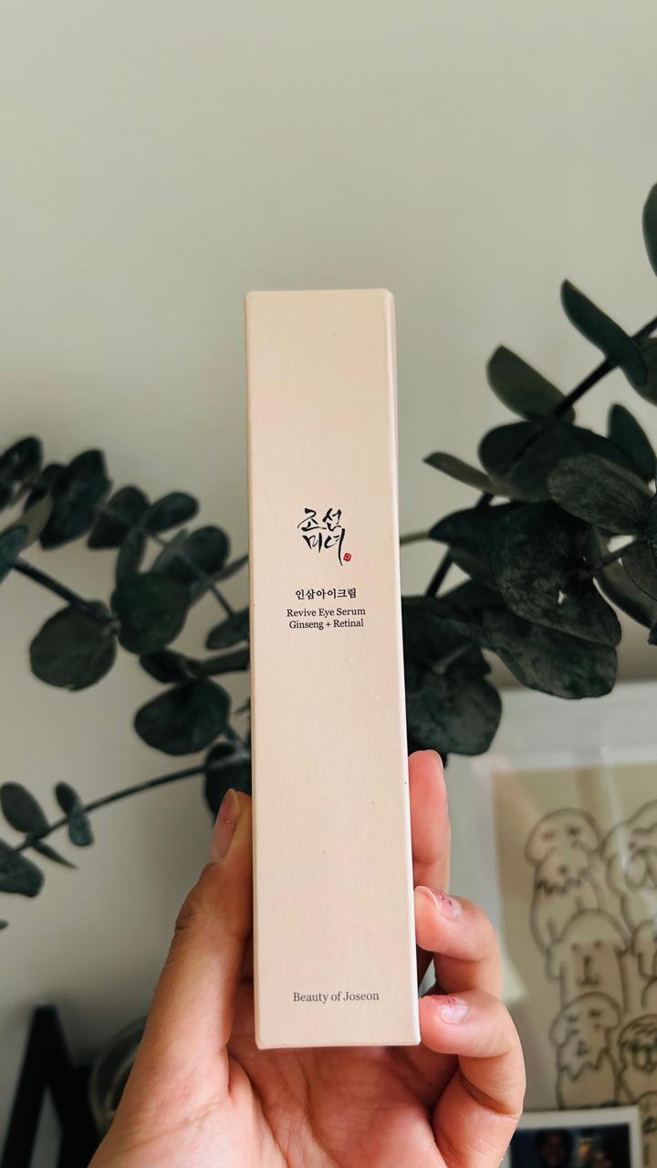 Una mano sostiene una caja beige etiquetada como Beauty of Joseon Revive Eye Serum Ginseng + Retinal de Bemba, resaltando su fórmula para el contorno de ojos con infusión de ginseng, con tallos de hojas verdes y un marco de fotos borroso en el fondo.
