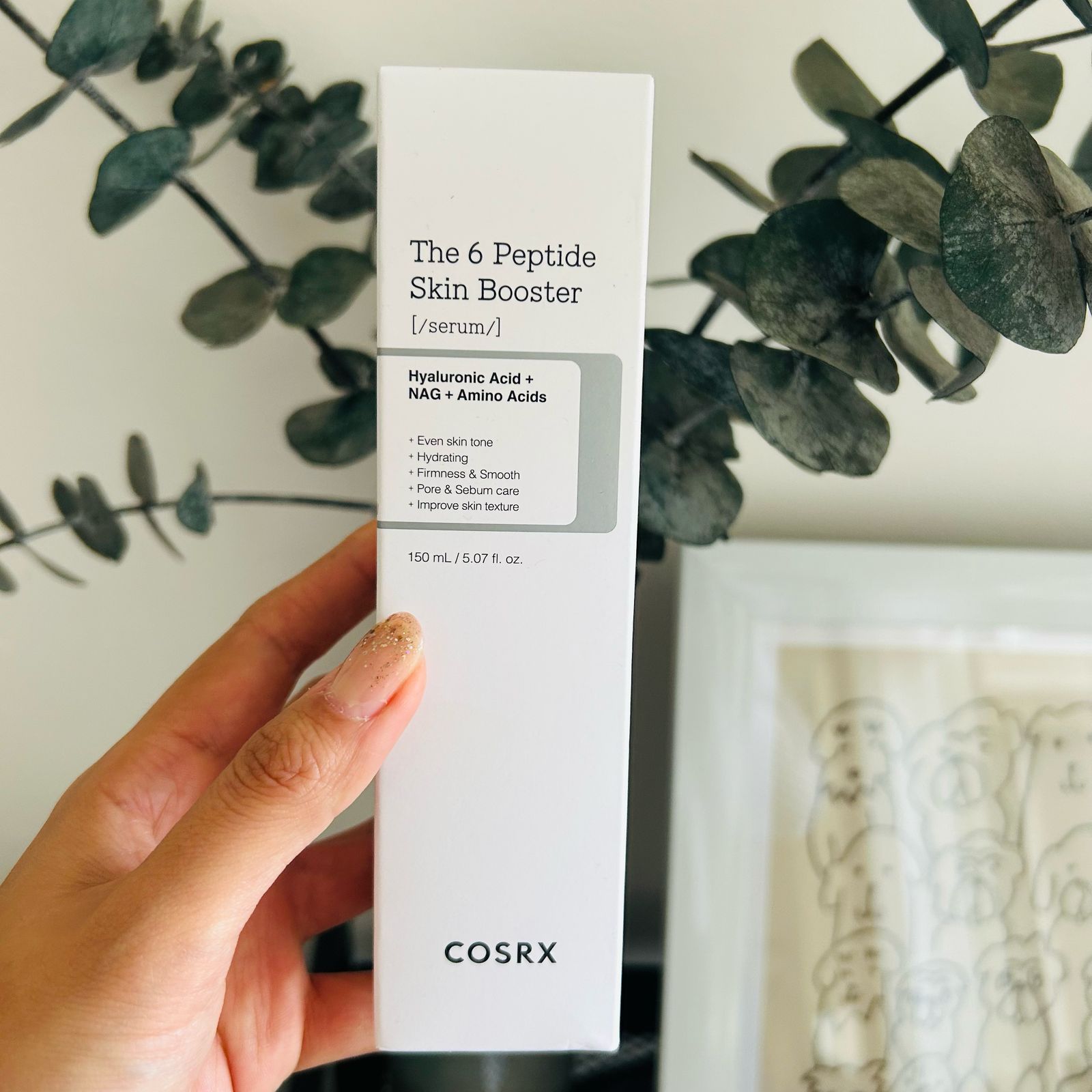 Una mano sostiene una caja blanca de Bemba. COSRX, el sérum potenciador de la piel de 6 péptidos, frente a ramas de plantas frondosas y un dibujo lineal enmarcado al fondo.