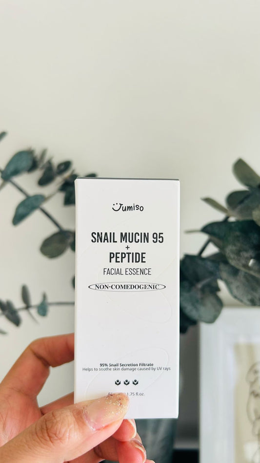 Una mano sostiene una caja blanca etiquetada como Bemba. Jumiso Snail Mucin 95 + Peptide Essence 100 ml, que destaca la mucina de caracol y los péptidos para aumentar la elasticidad de la piel, sobre un fondo difuminado con plantas verdes.