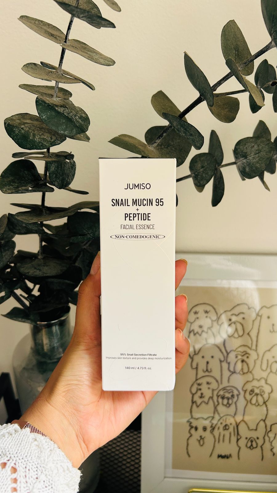 Una mano sostiene una caja de Bemba. Jumiso Esencia Facial con Mucina de Caracol 95% + Péptidos (140 ml), conocida por su mucina de caracol y sus péptidos, con hojas verdes y una ilustración de un perro enmarcada al fondo.