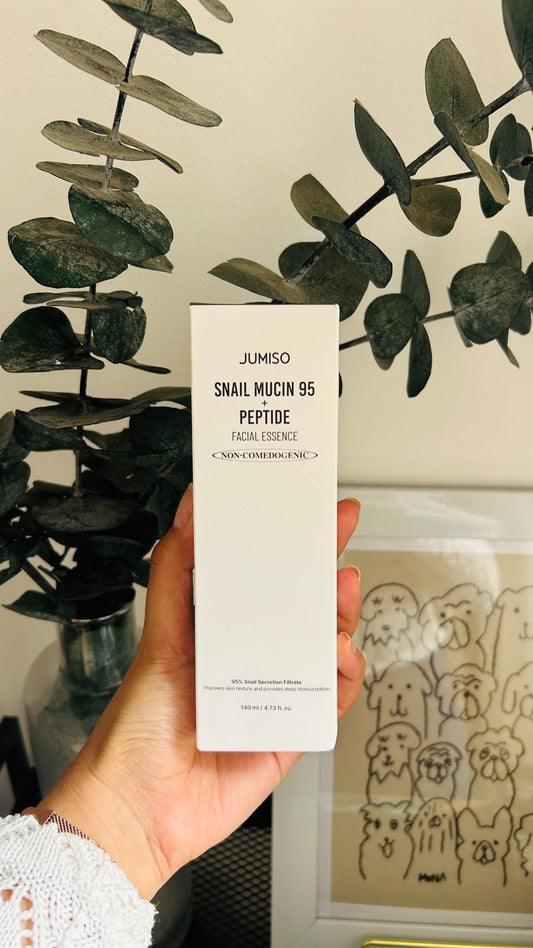 Una mano sostiene una caja de Bemba. Jumiso Esencia Facial con Mucina de Caracol 95% + Péptidos (140 ml), conocida por su mucina de caracol y sus péptidos, con hojas verdes y una ilustración de un perro enmarcada al fondo.