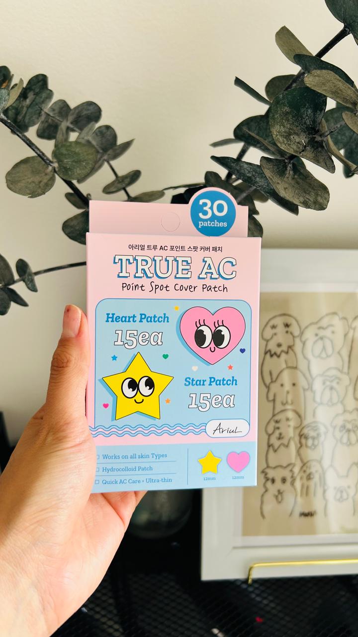 Una mano sostiene una caja de Bemba. Ariul True AC Point Spot Cover 30 parches hidrocoloides, con diseños de corazones y estrellas que ayudan a reducir la inflamación y proteger el acné. Al fondo, plantas y una ilustración marcada de perros.
