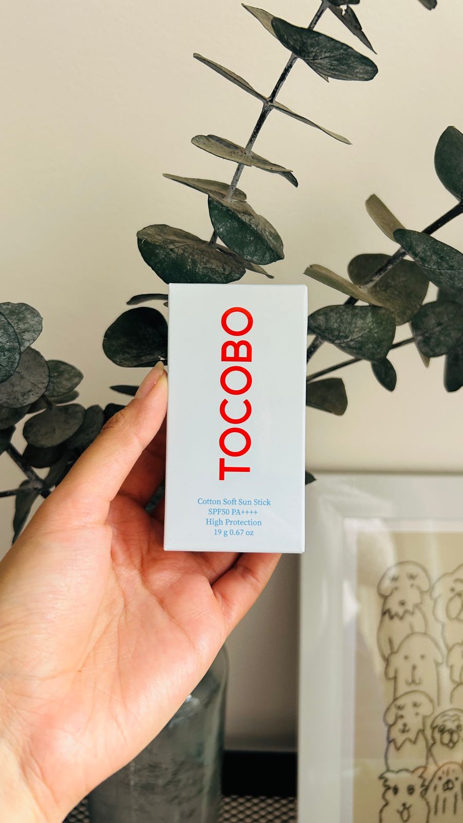 Una mano sostiene una caja blanca con la etiqueta Tocobo de Bemba, que dice "protección solar con acabado mate". La caja, colocada delante de hojas de eucalipto verde oscuro y una obra de arte enmarcada, dice "Cotton Soft Sun Stick High Protection".