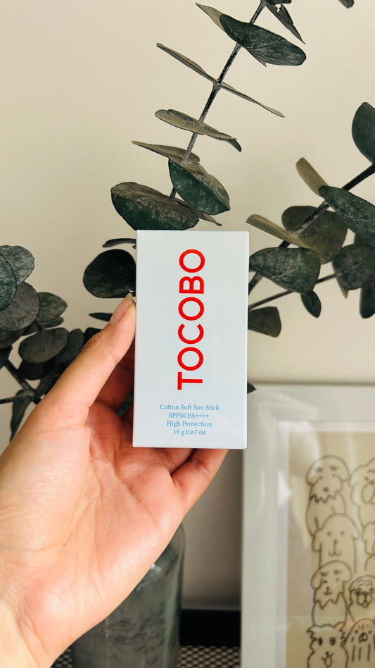Una mano sostiene una caja blanca con la etiqueta Tocobo de Bemba, que dice "protección solar con acabado mate". La caja, colocada delante de hojas de eucalipto verde oscuro y una obra de arte enmarcada, dice "Cotton Soft Sun Stick High Protection".