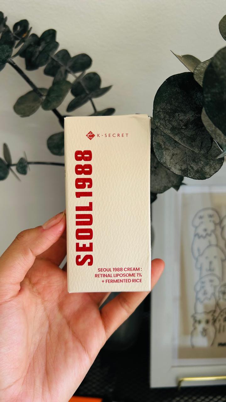 Una mano sostiene una caja color crema con la etiqueta "SEÚL 1988" en rojo. El texto dice "KSECRET - Suero SEÚL 1988: Liposoma Retinal 2% + Ginseng Negro" de Bemba. Al fondo aparecen hojas y un dibujo enmarcado.