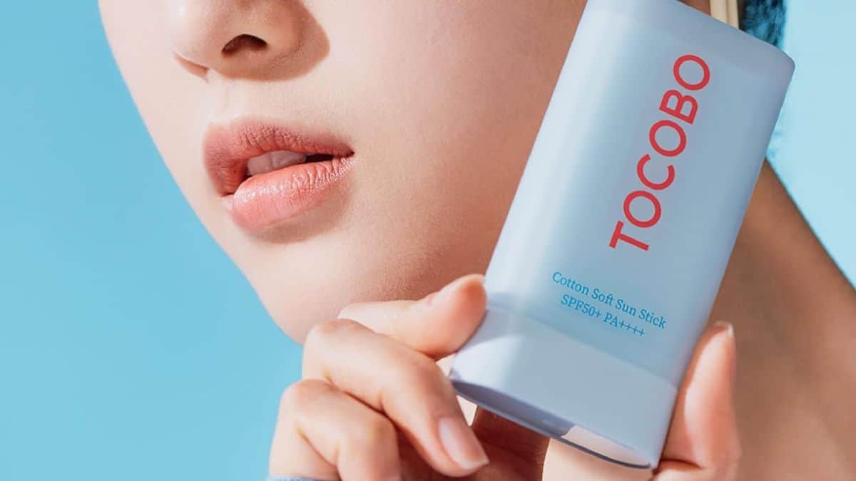 Una persona sostiene un protector solar Bemba. Tocobo Sun Stick FPS 50+ PA++++ azul claro cerca del rostro sobre un fondo azul, destacando la protección solar avanzada y las hierbas calmantes para un acabado mate perfecto.