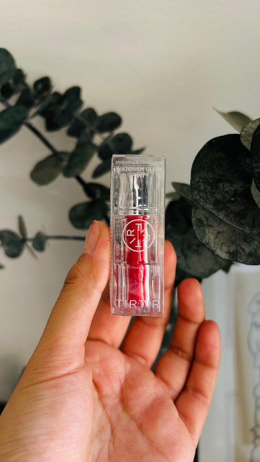 Una mano sostiene un tubo transparente de TIRTIR Waterism Glow Mini Tinta de Bemba., su vibrante tinte rojo ideal para labios secos, sobre un fondo borroso de follaje verde y paredes claras.