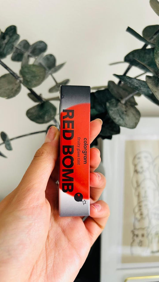 Una mano sostiene una caja rectangular etiquetada como Colorgram Fruity Glass Tint Red Bomb de Bemba, con un vibrante diseño en rojo y negro. Al fondo se ven hojas verdes y una obra de arte enmarcada y difuminada.