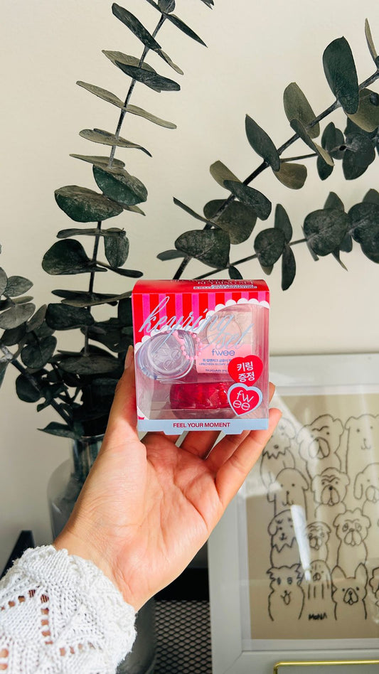 Una mano sostiene el Fwee Lip&Cheek Glowy Jelly Pot + Key Ring de Bemba, un bálsamo rojo con texto en coreano que promete brillo sin sensación pegajosa, frente a ramas de eucalipto y una ilustración enmarcada de un perro de dibujos animados.