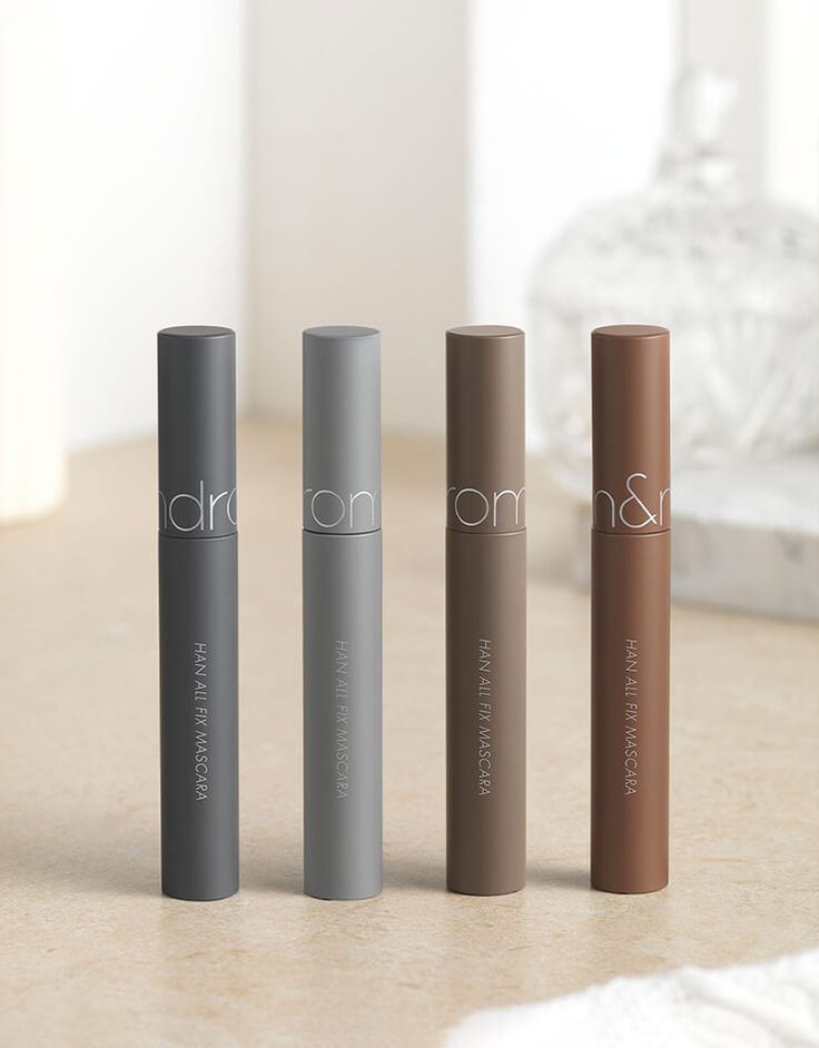 Cuatro tubos de Rom&nd Han All Fix Mascara de Bemba., en empaque mate, se alinean sobre una superficie beige. Los tonos son gris oscuro, gris claro, taupe y marrón, destacados sobre un fondo suavemente desenfocado.