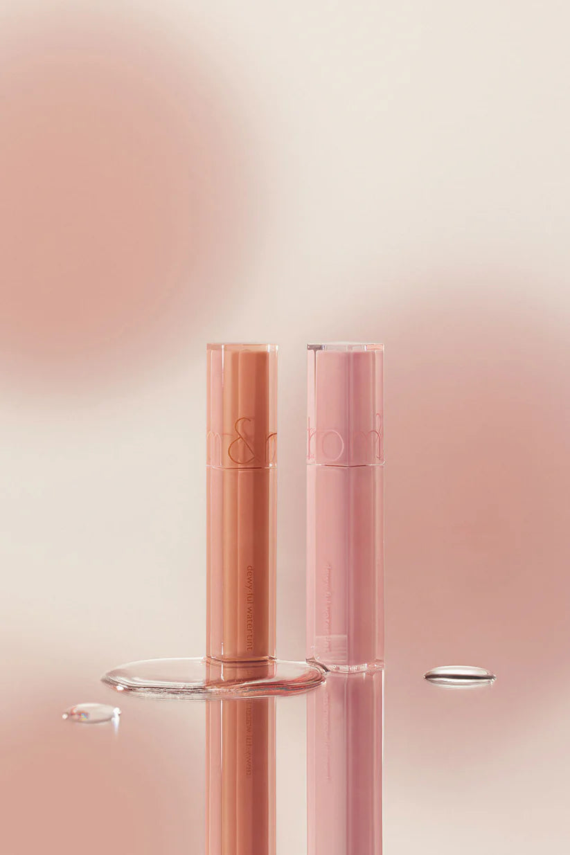 Dos labiales Rom&nd Dewyful Water Tint de Bemba —uno melocotón y otro rosa claro— se yerguen sobre una superficie reflectante con gotas de agua. Un suave fondo beige con degradados rosas realza su acabado brillante y larga duración.
